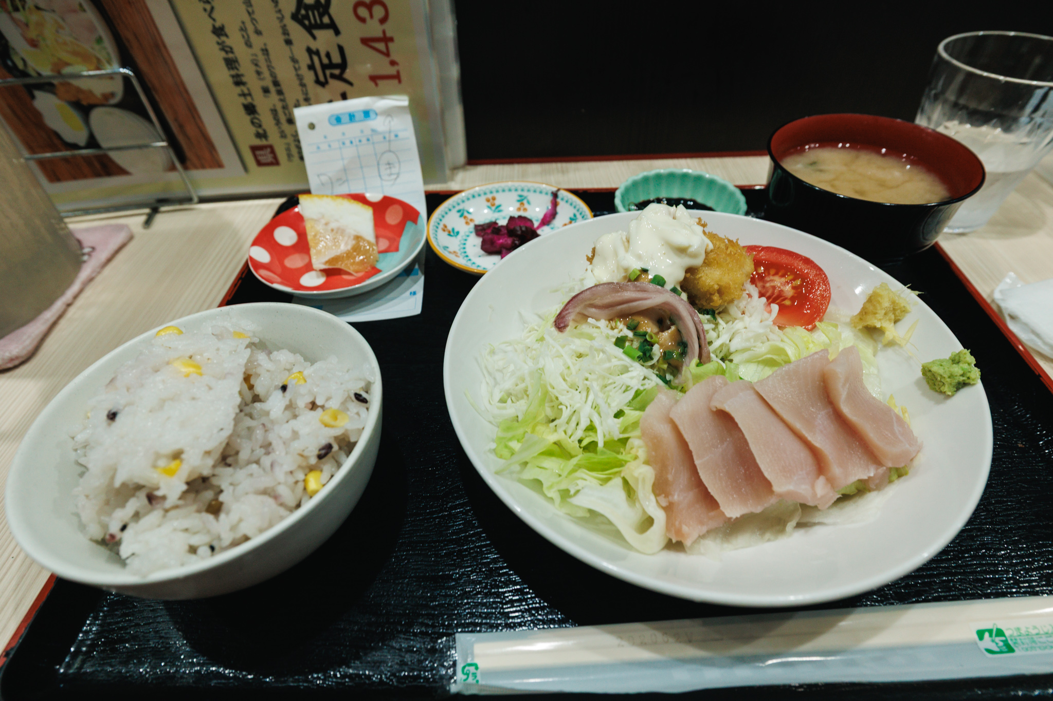 The wani set meal on a black tray: rice with corn and grains, miso soup, purple pickles, a slice of citrus, and a large plate of shark sashimi, fried shark under tartar sauce and vinegared shark with salad, beside a menu stand and a water glass