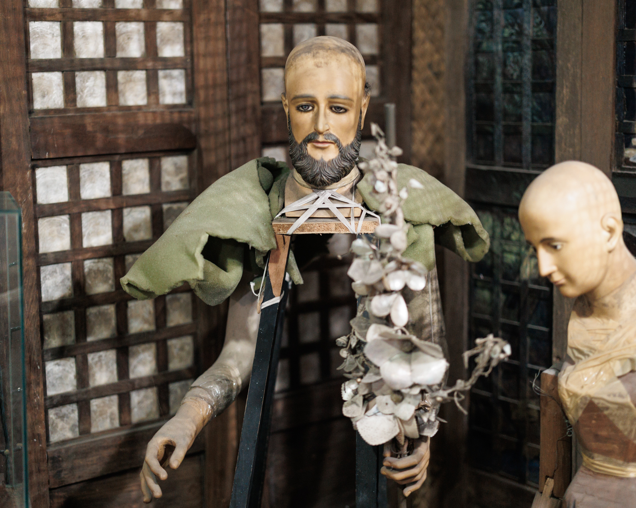 A carved saint with a bearded face and painted hands, a worn green cloth over his shoulders and a bare wooden post where his body would be, in front of lattice windows of pale translucent squares, with another bald figure beside him